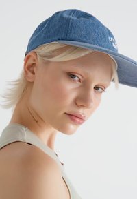 Boné de denim com aba curva, apresentando um logótipo na parte da frente. Cor azul clara com um acabamento texturizado. Acompanhado por uma t-shirt sem mangas cinza.