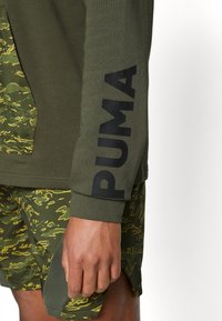 Olivgrüner Sweatshirt mit schwarzem "PUMA"-Logo am Ärmel, kombiniert mit grünen Shorts, die ein wellenförmiges gelbes Muster und eine strukturierte Seitenpartie aufweisen.