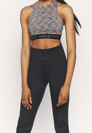 Vrouw die een grijs Nike Pro mouwloos crop top draagt en zwarte, hooggetailleerde leggings, staand met haar armen ontspannen langs haar zij.