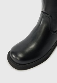 Botă din piele neagră cu vârful rotunjit, textură netedă și talpă groasă din cauciuc. Prezintă cusături minime și fără elemente metalice vizibile.