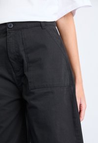 Schwarze weit geschnittene Hosen aus Baumwolle mit einem Knopfverschluss, seitlichen Taschen und einer glatten Textur. Nahaufnahme, die den Bund und den Stoff in den Fokus rückt.