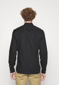 Persona con capelli ricci che indossa una maglietta nera a maniche lunghe e pantaloni beige, vista da dietro su uno sfondo bianco.