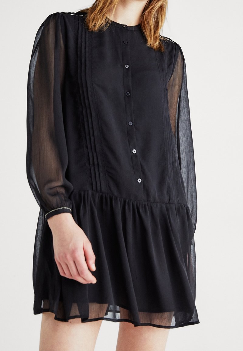Femme portant une robe longue noire transparente à manches longues avec des boutons sur le devant, des détails aux épaules et un ourlet de jupe froncé sur un fond uni.
