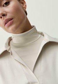 Moteris vilki aukštai apykakle bežinę palaidinę, užsidėjusi šviesiai kreminė marškiniais, prieš paprastą šviesią foną.