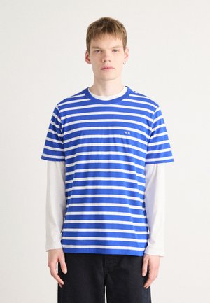 Camiseta de rayas azules y blancas con mangas cortas, escote redondo y pequeño logotipo bordado "MN" en el pecho. Superpuesta sobre una camisa de manga larga blanca.