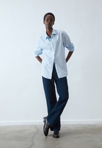 Chemise bleu clair à manches retroussées, associée à un jean large bleu foncé. Des chaussures noires complètent la tenue. Style épuré et minimaliste.