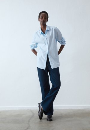 Camicia button-up blu chiaro con maniche arrotolate, abbinata a pantaloni in denim a gamba larga blu scuro. Scarpe nere completano l'outfit. Stile pulito e minimalista.