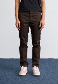Brown fitted trousers with a smooth texture, paired with pastel pink sneakers featuring white laces and a textured sole.