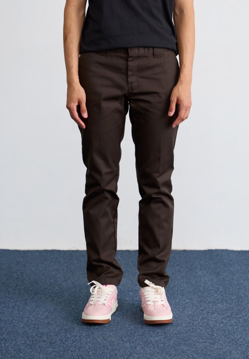 Brown fitted trousers with a smooth texture, paired with pastel pink sneakers featuring white laces and a textured sole.
