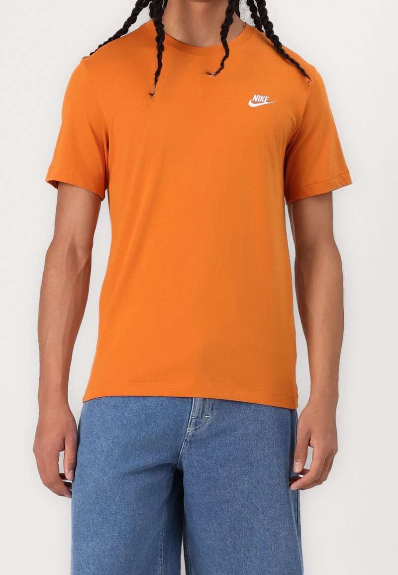 Oranžové tričko s krátkým rukávem vyrobené z bavlny, s bílým logem Nike na levé straně hrudi. Nosené s modrými denimovými šortkami.