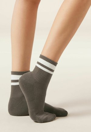 Pair of gray ankle socks with two white stripes worn on feet with one heel lifted against a plain light background.