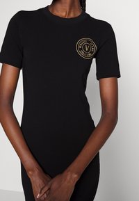 Sort kortærmet tætsiddende kjole med gyldigt rundt 'Versace Jeans Couture' logo på venstre bryst båret af person med foldede hænder.