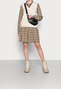 Pull en V beige porté sur une robe à carreaux bruns avec des boutons, associé à des bottes montantes beiges. Un sac à bandoulière noir avec des détails en chaîne.