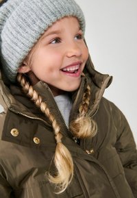 Veste matelassée marron avec col haut, boutons-pression dorés et fermeture éclair. Bonnet en tricot côtelé gris clair. Cheveux blonds en tresses.
