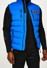 Blauer Stepp-Weste mit hohem Kragen, zwei Reißverschlusstaschen und einem Logopatch. Hergestellt aus glänzendem, leichtem Stoff mit gestepptem Muster.