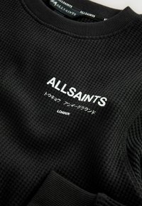 Čierny, textúrovaný sveter s okrúhlym výstrihom. Na prednej strane je biely logotyp "ALLSAINTS" s japonským textom a pod ním nápis "LONDON".