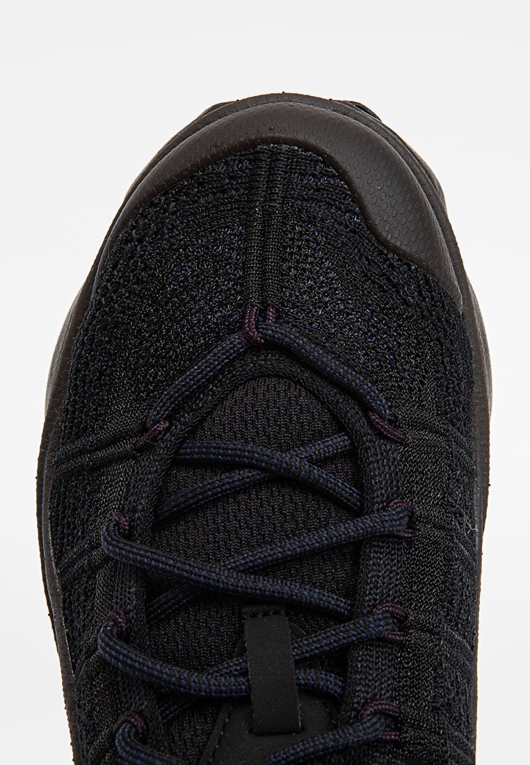 Botte de sport noire avec un dessus en tricot texturé, accents foncés et lacets visibles. Elle présente une pointe arrondie et une semelle en caoutchouc flexible.