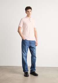 Ρόδινο πόλο μπλουζάκι με γιακά και τρία κουμπιά, συνδυασμένο με μπλε τζιν παντελόνι. Το μοντέλο φορά μαύρες γαλότσες. Λείο ύφασμα, άνετη εφαρμογή.