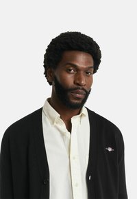 Schwarze Strickjacke mit Knopfverschluss über einem weißen Hemd mit Kragen, mit einem kleinen gestickten Logo auf der linken Seite. Die Textur wirkt weich.