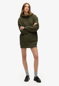 Robe à capuche vert olive avec une poche avant, un cordon de serrage et des poignets côtelés. Associée à des chaussettes grises et des chaussures noires.