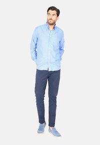 Camicia a maniche lunghe di colore blu chiaro con piccoli motivi bianchi, pantaloni blu scuro e mocassini in suede blu. Il modello è in piedi con le mani in tasca.