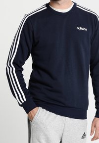 Sudadera azul marino con diseño de tres franjas blancas en las mangas y pequeño logo blanco en el pecho. Puños y bajo de canalé. Fabricada con material suave.