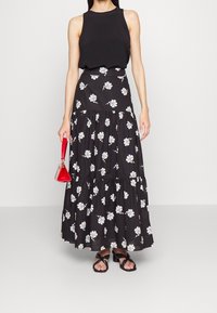 Falda larga negra con estampado de flores blancas, diseño en capas, combinada con una blusa sin mangas negra y un bolso rojo. Sandalias con tiras.