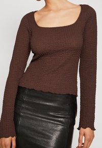 Brun strukturerad långärmad topp med fyrkantig halsringning och vågiga kanter, ihop med en högmidjad svart läderkjol.