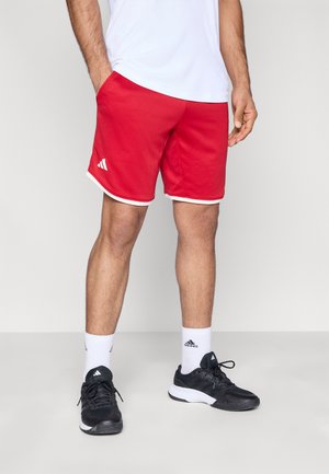 Rote Sportshorts mit weißer Einfassung aus strukturiertem Stoff, mit Adidas-Logo an der Seite. Kombiniert mit schwarzen Schuhen und weißen Socken.