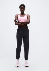 Άτομο με αθλητική εμφάνιση στέκεται με αυτοπεποίθηση με τα χέρια σταυρωμένα, φορώντας ροζ αθλητικό σουτιέν, μαύρα παντελόνια τζόγκινγκ και ροζ αθλητικά παπούτσια σε απλό φόντο.