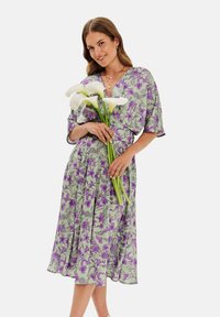Kvetinové šaty s obalom v svetlozelené farbe s fialovými a čiernymi botanickými vzormi. Krátke rukávy, výstrih do V a naťahovaný pás, doplnené o biele kalá. 