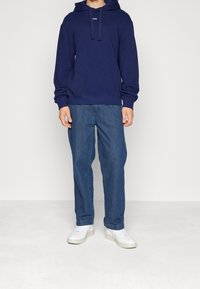 Námořnická modrá mikina s logem na přední straně, spárovaná s volnými světle modrými džíny a bílými teniskami s texturovaným povrchem.