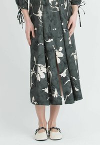Grünes, blumengeprägtes Kleid mit langen Ärmeln und Schlitzen, das eine glatte Textur aufweist. Getragen mit cremefarbenen Schuhen mit schwarzen Riemen.