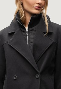 Svart dubbelknäppt kappa i ullblandning, med ett vadderat innerfoder, hög krage och framsidan med dragkedjedetalj och knappar.