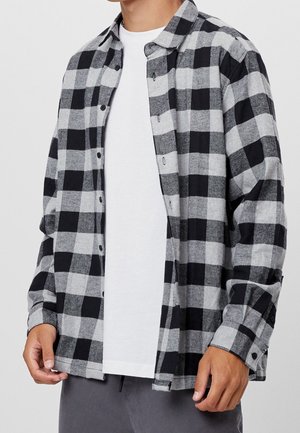 Mężczyzna noszący rozpiętą czarno-szarą flanelową koszulę w kratę na gładkim białym t-shircie i szarych spodniach.