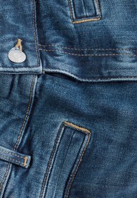 Denimstof in medium blauw met vervaagde textuur. Bevat een zilveren knoop en contrasterende gouden stiksel, met zichtbare zak- en naaddetails.