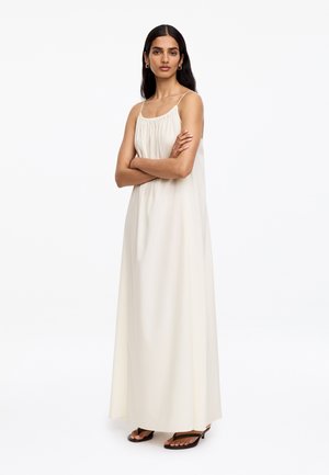 Femme aux longs cheveux noirs, debout les bras croisés, portant une robe maxi sans manches crème et des sandales noires à talons.