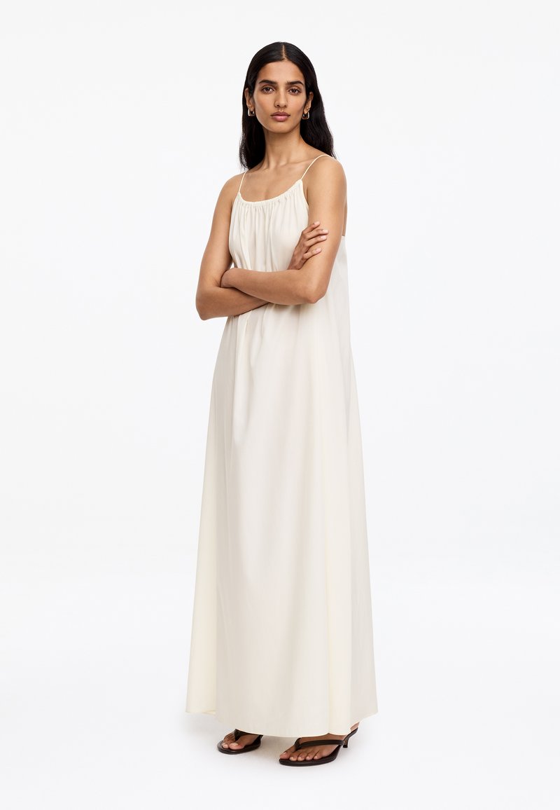 Femme aux longs cheveux noirs, debout les bras croisés, portant une robe maxi sans manches crème et des sandales noires à talons.