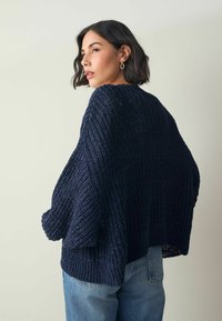 Žena s krátkými tmavými vlasy, která má na sobě zlaté kruhové náušnice, tmavomodrý silný pletený svetr a světle modré džíny, stojí bokem na neutrálním pozadí.