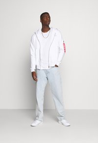 Haut de zip blanc avec une étiquette rouge, porté sur un t-shirt blanc, associé à un jean bleu clair ample et des baskets blanches.