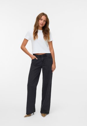 T-shirt blanc court associé à un jean noir à taille haute et jambes larges. Le modèle porte des escarpins pointus à imprimé serpent et arbore des cheveux doux et ondulés.