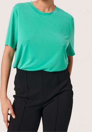 Osoba ubrana w luźną, turkusową bluzkę z krótkim rękawem wpuszczoną w wysokowciskie, dopasowane czarne spodnie, stojąca z jedną ręką swobodnie opuszczoną.