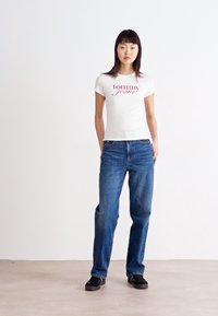 Balta prigludusi marškinėliai su raudonu "tommy jeans" logotipu, derinami su mėlynomis džinsomis. Modelis vilki juodus batus. Paprasta fono atmosfera ir neutrali spalvų paletė.