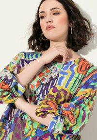 Värikäs, kuviollinen paita löysällä istuvuudella, jossa on eloisia vihreitä, sinisiä, purppuroita ja oransseja sävyjä. Lyhyet hihat sidottuna hiha-aukkoihin.