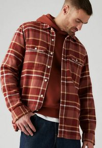 Camicia di flanella a quadri rossa e beige con tasche frontali e bottoni bianchi, indossata sopra un hoodie marrone e jeans scuri. Tessuto texture.