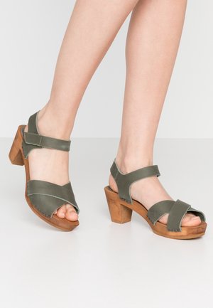 Füße in grünen Ledersandalen mit Holzblockabsätzen und Knöchelriemen auf weißem Boden vor schlichter Kulisse.