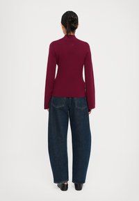 Sötét hajú nő kontyban, testhezálló bordó hosszú ujjú felsőt, széles lábú sötétkék farmernadrágot és fekete lapos cipőt visel, háttal áll.