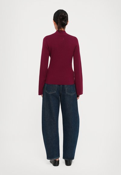 Kvinne med mørkt hår i en knute, iført en tettsittende burgunder langermet topp, vide mørkeblå jeans og svarte flate sko, som står med ryggen til.
