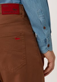 Pantaloni di cotone marrone con una toppa rossa con logo sulla tasca posteriore, abbinati a una camicia di denim azzurro con bottoni marroni.