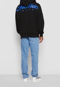 Sudadera con capucha negra con impresión azul de "Calvin Klein Jeans" en la espalda, combinada con vaqueros de mezclilla azul claro y zapatillas blancas con detalles de logotipo.